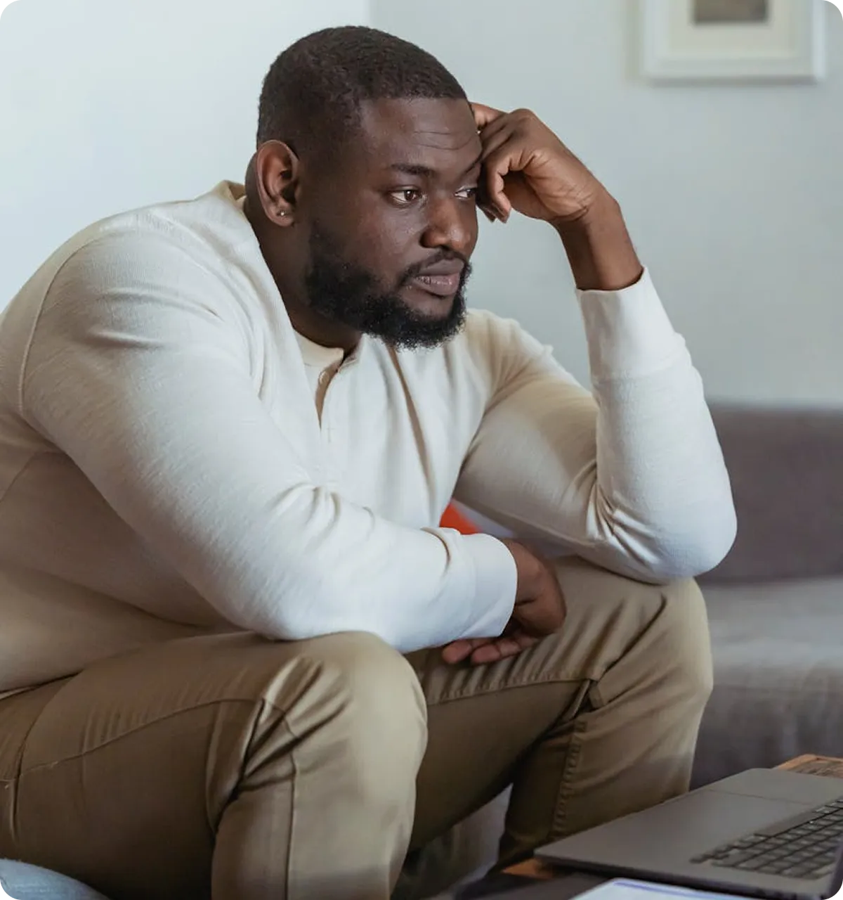 Black man looking thoughtful while sitting on couch with laptop - depression therapy and mental health counseling in Virgi...